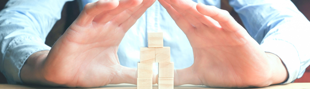 Hände des Managements werden schützend über ein Haus aus Holzwürfeln gehalten